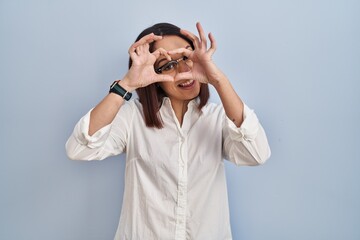 Young hispanic woman standing over white background doing heart shape with hand and fingers smiling looking through sign