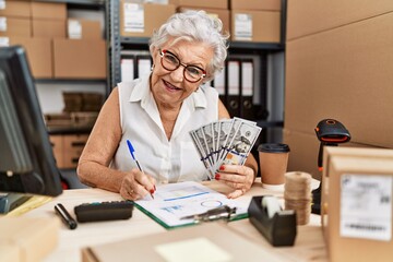 Senior grey-haired woman business worker holding dollars and writing on checklist at office