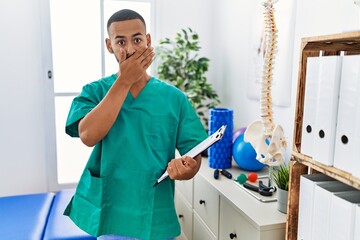 African american physiotherapist man working at pain recovery clinic shocked covering mouth with hands for mistake. secret concept.