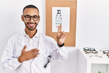 African american optician man standing by eyesight test smiling swearing with hand on chest and...