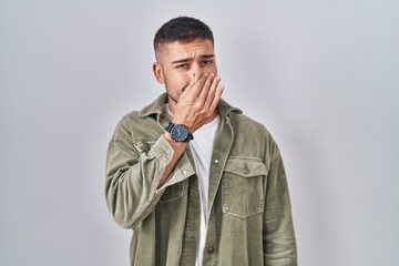 Young hispanic man standing over isolated background smelling something stinky and disgusting,...