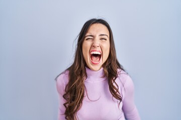 Young brunette woman standing over blue background angry and mad screaming frustrated and furious, shouting with anger. rage and aggressive concept.
