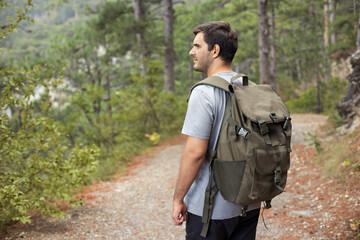 View from the back of young man with a hiking backpack walks along a path through a forest in the mountains. Active and healthy lifestyle concept.