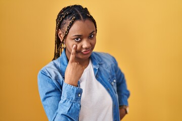 African american woman with braids standing over yellow background pointing to the eye watching you gesture, suspicious expression