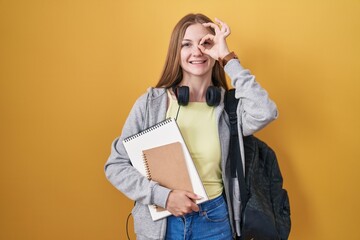 Young caucasian woman wearing student backpack and holding books doing ok gesture with hand smiling, eye looking through fingers with happy face.