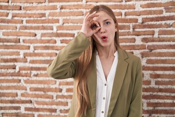 Young caucasian woman standing over bricks wall background doing ok gesture shocked with surprised face, eye looking through fingers. unbelieving expression.