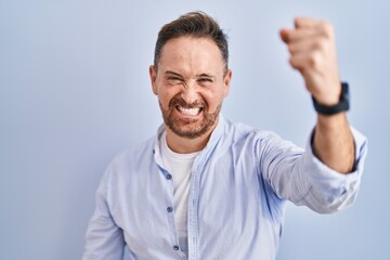 Middle age caucasian man standing over blue background angry and mad raising fist frustrated and furious while shouting with anger. rage and aggressive concept.