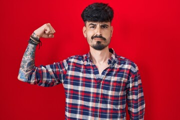 Young hispanic man with beard standing over red background strong person showing arm muscle, confident and proud of power
