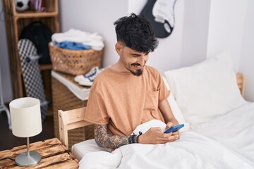 Young hispanic man using smartphone sitting on bed at bedroom