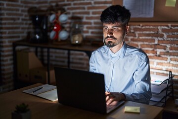 Young hispanic man with beard working at the office at night relaxed with serious expression on face. simple and natural looking at the camera.