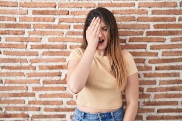 Young brunette woman standing over bricks wall yawning tired covering half face, eye and mouth with hand. face hurts in pain.