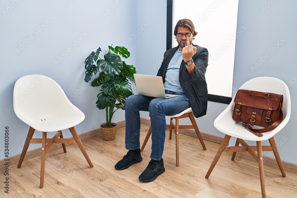 Wall mural Handsome middle age man sitting at waiting room working with laptop showing middle finger, impolite and rude fuck off expression