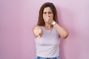 Brunette woman standing over pink background laughing at you, pointing finger to the camera with...