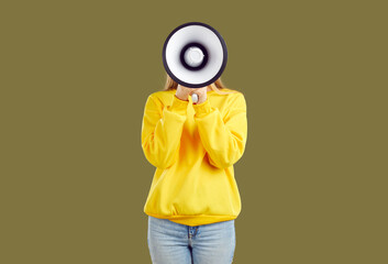 Young woman makes loud announcement hiding her face behind megaphone on khaki background in studio....