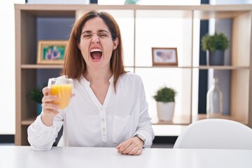Brunette woman drinking glass of orange juice angry and mad screaming frustrated and furious, shouting with anger. rage and aggressive concept.