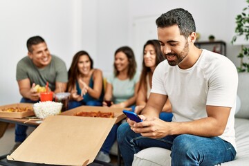 Group of young hispanic friends eating italian pizza sitting on the sofa. Man smiling happy and using smartphone at home.