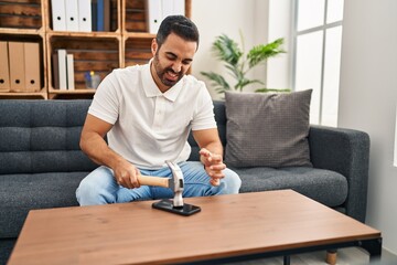 Young hispanic man stressed breaking smartphone with hammer at home