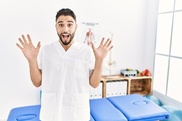 Young handsome man with beard working at pain recovery clinic celebrating crazy and amazed for success with arms raised and open eyes screaming excited. winner concept