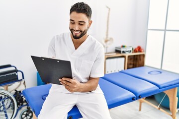 Young arab man wearing physiotherapist uniform writing on clipboard at clinic