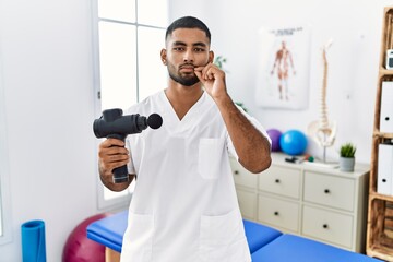 Young indian physiotherapist holding therapy massage gun at wellness center mouth and lips shut as...