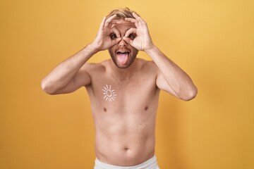 Fototapeta premium Caucasian man standing shirtless wearing sun screen doing ok gesture like binoculars sticking tongue out, eyes looking through fingers. crazy expression.