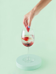 Young woman's hand puts strawberry in glass with infused detox water with fresh red berries. Isolated pastel green background. Minimal concept of summer party healthy beverages. Creative card.