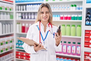 Young blonde woman working at pharmacy drugstore holding bottle winking looking at the camera with...