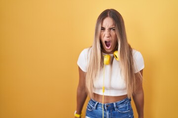 Young blonde woman standing over yellow background wearing headphones angry and mad screaming...