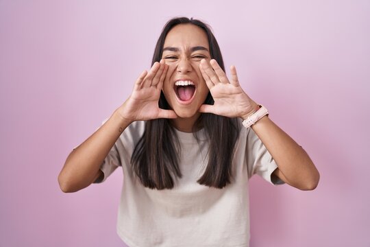 Young hispanic woman standing over pink background shouting angry out loud with hands over mouth