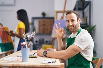 Fototapeta premium Hispanic man at painter studio doing ok sign with fingers, smiling friendly gesturing excellent symbol