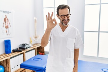 Middle age man with beard working at pain recovery clinic smiling positive doing ok sign with hand and fingers. successful expression.