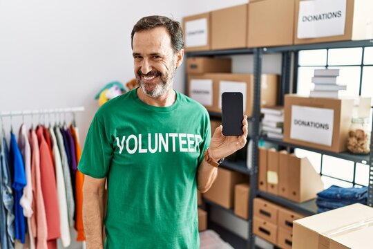 Middle age man with beard wearing volunteer t shirt holding smartphone looking away to side with smile on face, natural expression. laughing confident.