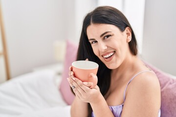 Young hispanic woman drinking cup of coffee sitting on bed at bedroom