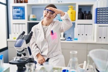 Hispanic girl with down syndrome working at scientist laboratory very happy and smiling looking far away with hand over head. searching concept.