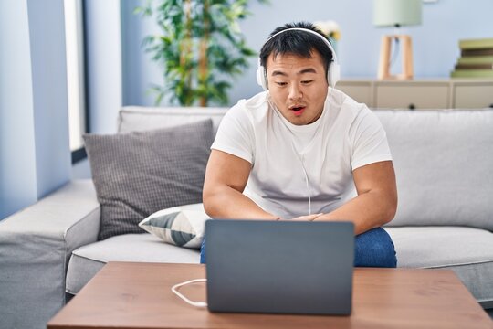 Young chinese man using laptop at home scared and amazed with open mouth for surprise, disbelief face