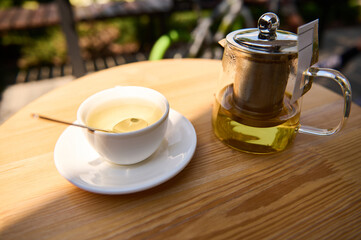 Close-up of a glass teapot and white cup with healthy green herbal tea, on a saucer, on a wooden table in the summer terrace. Food and drink consumerism