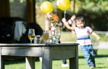 Glasses with drinks on a vintage table during a party. Happy children running with yellow balloons on blurred background