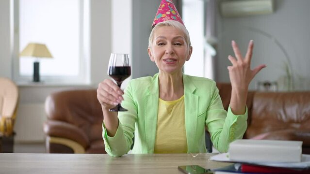 Portrait of confident woman toasting drinking red wine in slow motion. Beautiful Caucasian businesswoman in party hat celebrating holiday online from home office. Remote working lifestyle