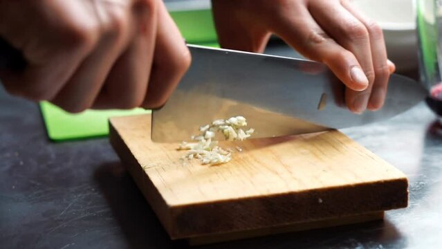 Horizontal close-up video of a knife chopping garlic with a knife on a board.