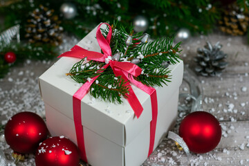 A close-up photo of a white gift box that stands on a wooden table among Christmas tree toys and coniferous branches.