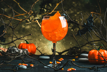 Halloween orange alcoholic cocktail on scary gray background with twisted branches, bats, stones,...