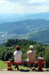 Fototapeta na wymiar A couple of tourists rest on a wooden bench on the top of Mount Zar. Toutists admire the view of lake Żwiecieckie. Beskids Mountains, Poland