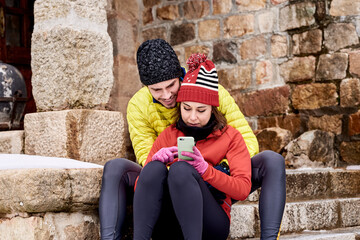 Couple in winter jackets and hats using cell phone sitting on stone steps.