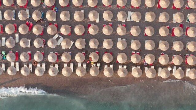 Beach with rows of thatch umbrellas - tiki straw parasols by the sea, waves crashing on the sand. Tourism on the Mediterranean. Aerial drone top down view on the coast at a summer vacation resort.