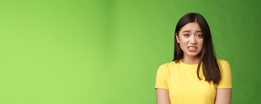 Anxious worried cute asian girl brunette clench teeth frowning shaking from fear look guilty afraid, stand nervous awaiting punishment, green background, apologizing ashamed