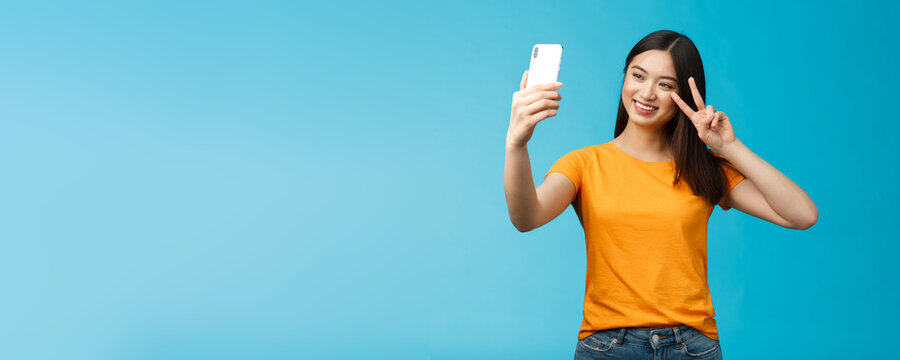 Friendly cheerful pretty asian woman brunette short haircut taking selfie, smiling joyfully hold smartphone show peace, victory sign at phone screen front camera, talking family video-call