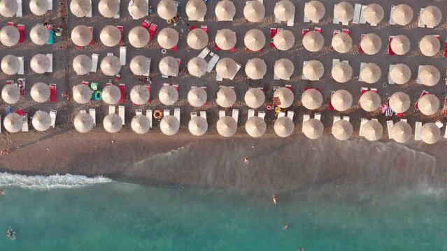Beach with rows of thatch umbrellas - tiki straw parasols by the sea, waves crashing on the sand. Tourism on the Mediterranean. Aerial drone top down view on the coast at a summer vacation resort.