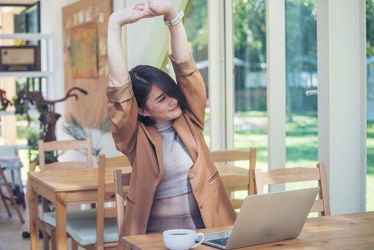 Woman stretching arm raised sitting incorrect position at home office desk. Young asian woman tired from work body stress back pain office syndrome. freelance Female work from anywhere using laptop