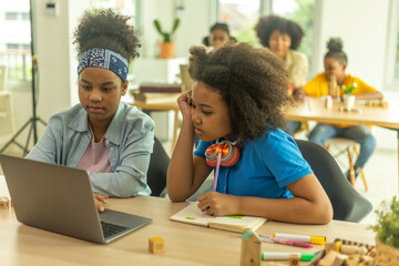 Two young African girls use computer notebook for an online class with support from a teacher. Kids...