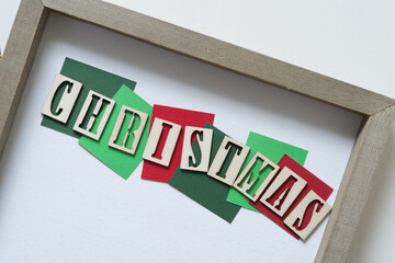 the word Christmas in wood stencil type on paper tiles arranged inside a wooden frame on blank paper
